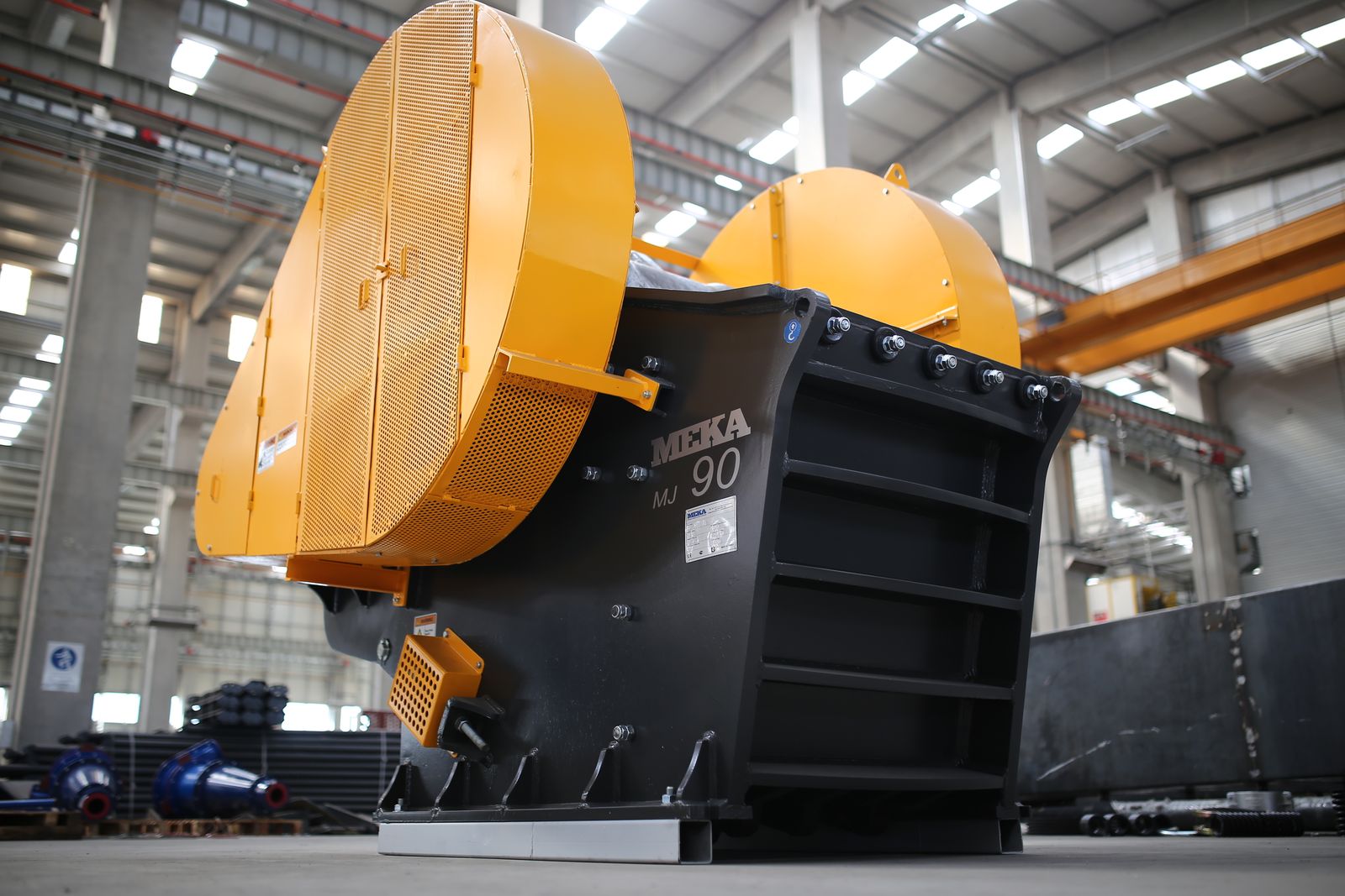 A yellow and black industrial jaw crusher photographed from below inside a factory hall, its heavy steel jaw and drive wheel dominating the frame.