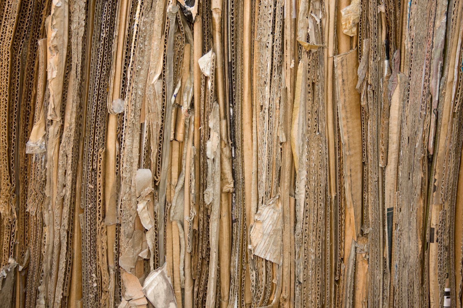 A tightly packed wall of flattened cardboard sheets seen head on, corrugated edges and torn fragments visible, filling the whole frame in shades of brown and cream