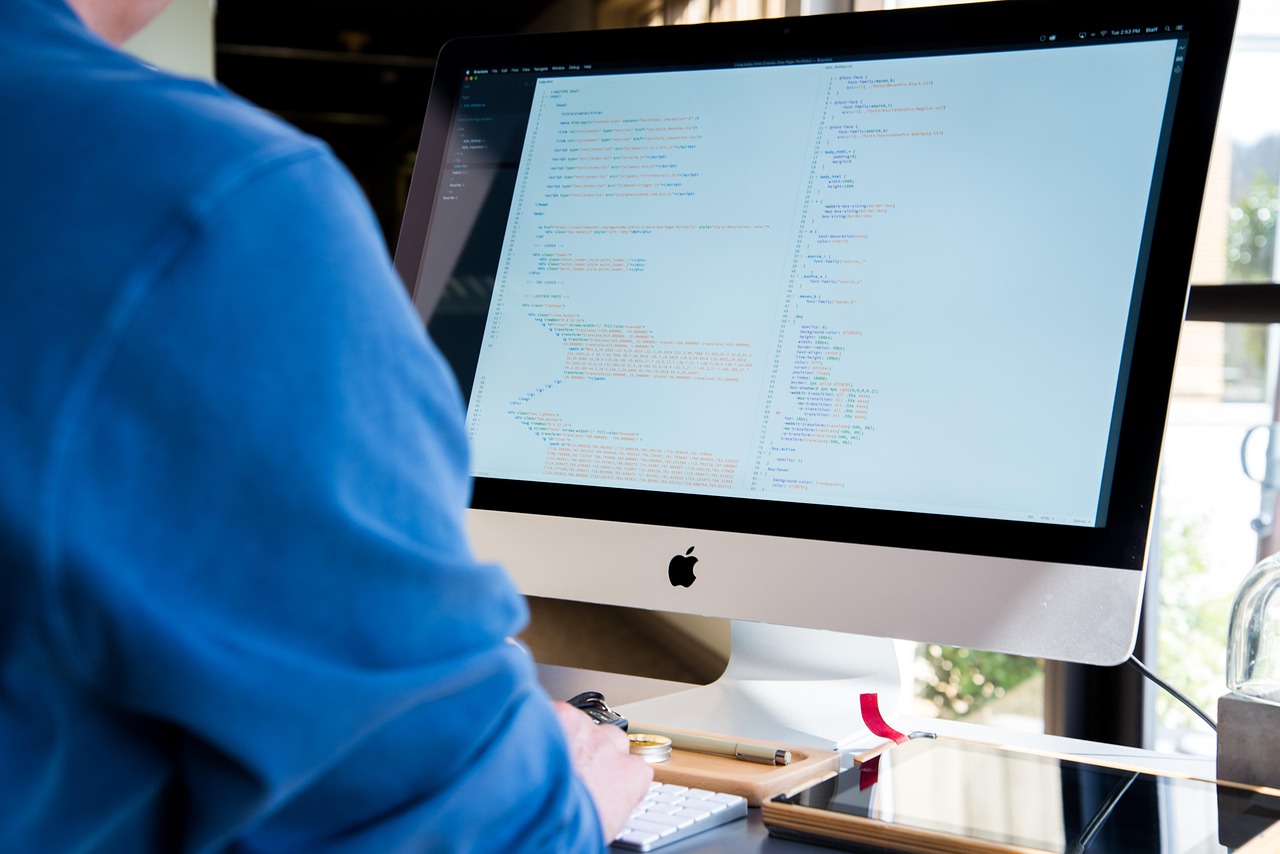 A person in a blue sweater working on an iMac that displays HTML and CSS source code.