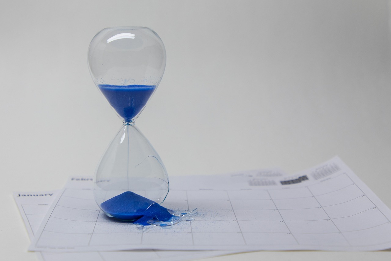 Eine Sanduhr mit zerbrochenem unterem Glaskolben steht auf Kalenderblättern, der blaue Sand ist ausgelaufen
