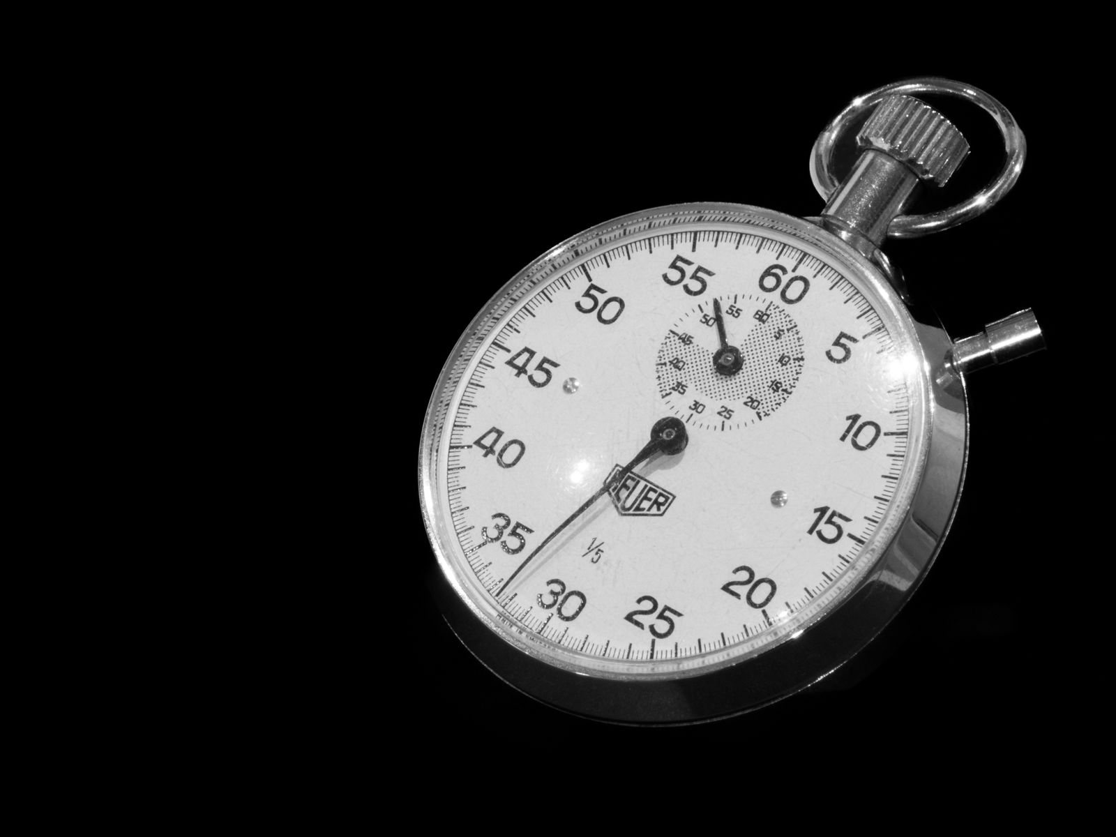 A chrome mechanical stopwatch photographed against a black background, its white dial marked in five second increments with a small sub dial for minutes, the crown and ring at the top right